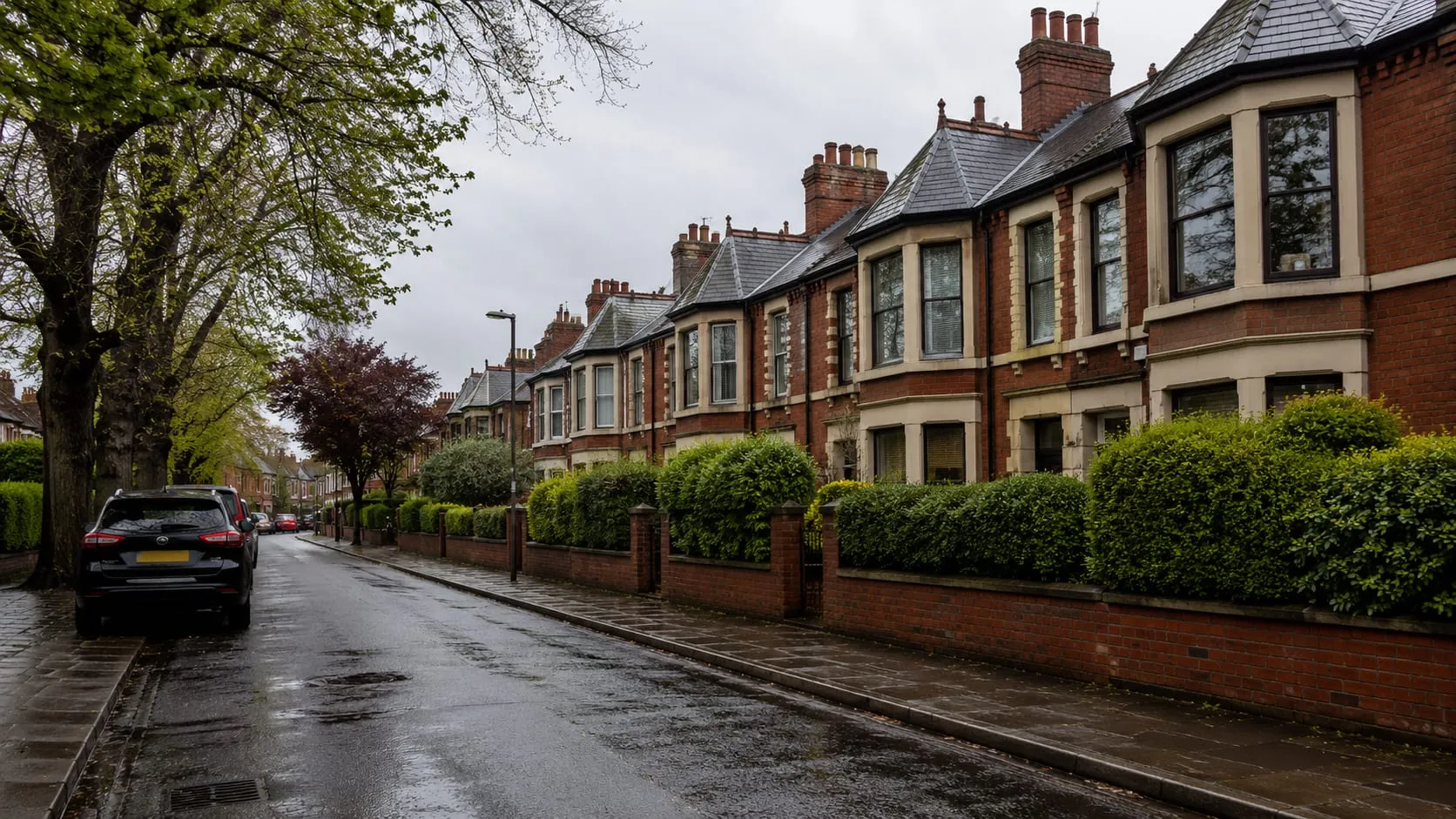 A residential street of red-brick Victorian terraced houses in south Manchester on a damp overcast day, cars parked along the kerb.